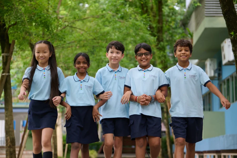 Five smiling children in blue uniforms walk arm-in-arm outdoors, surrounded by green trees, embodying friendship and joy.