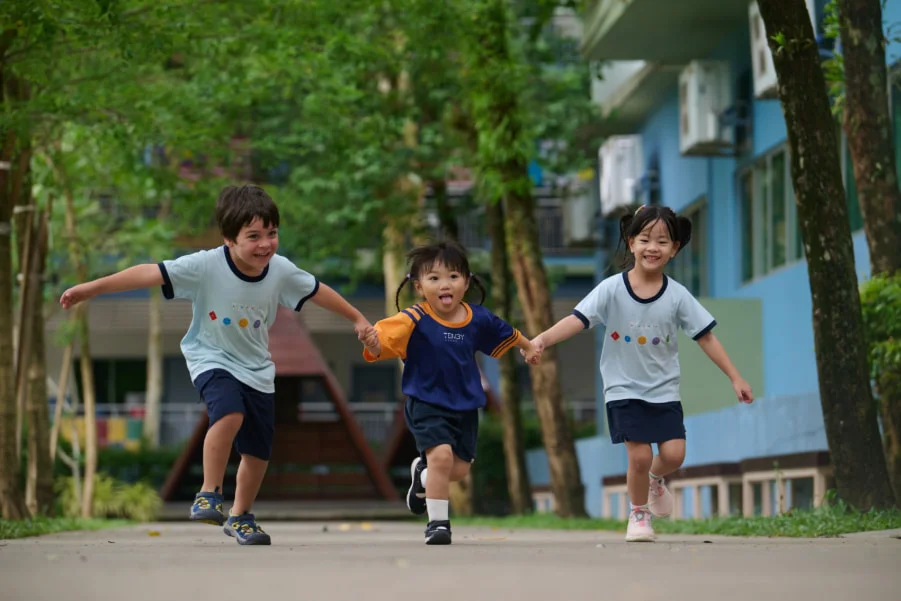 Three children joyfully run hand in hand down a tree-lined path, smiling and playing together on a sunny day.