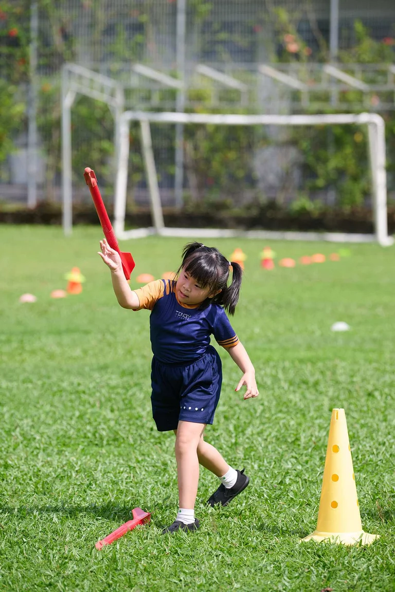 Young girl in sportswear throws red baton on grassy field, surrounded by cones, with a soccer goal in the background.