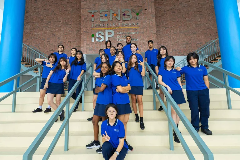 A group of students in blue uniforms pose on stairs at Tenby Schools Penang, under the International School Partnership sign.