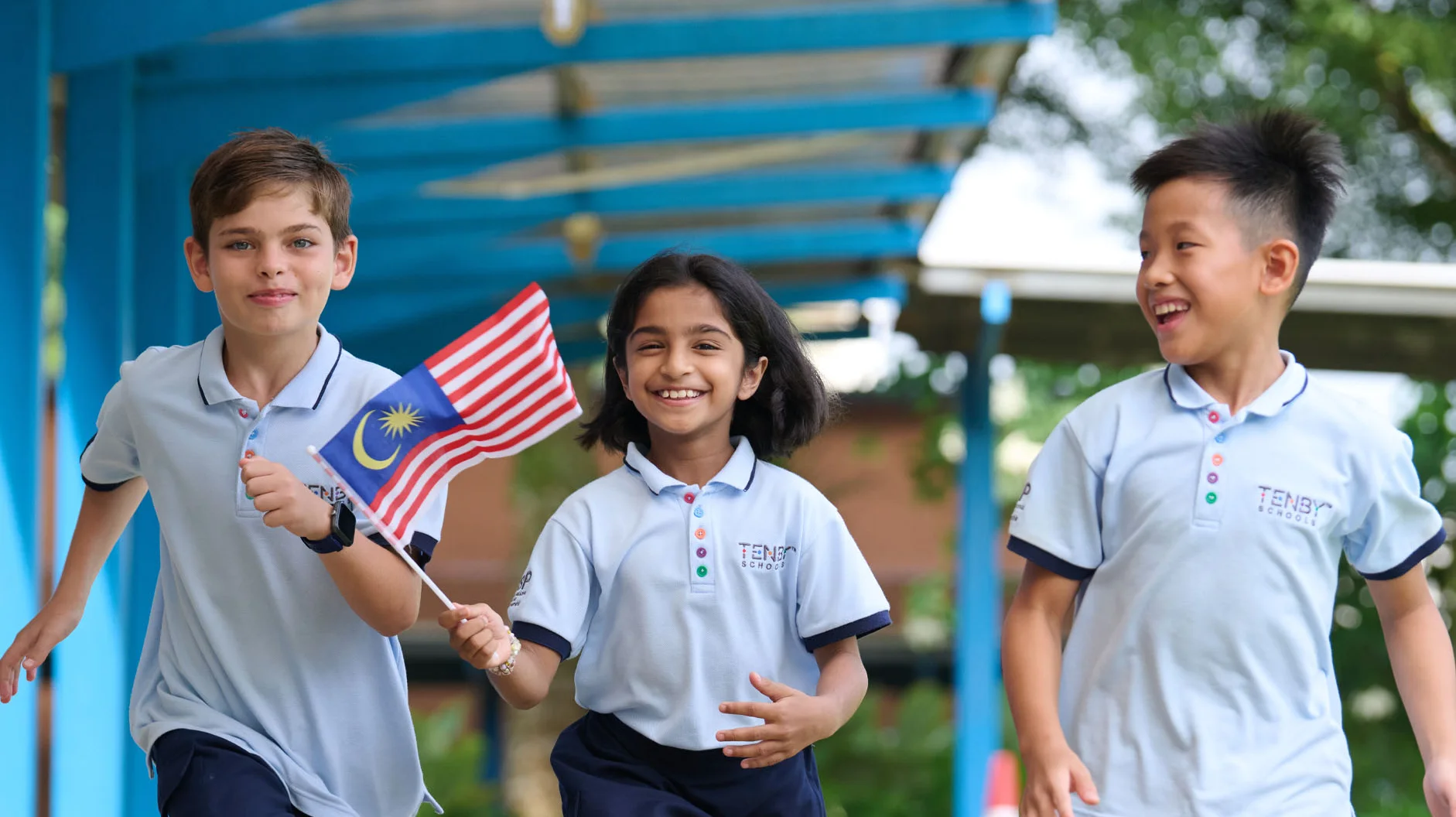 Three children in school uniforms joyfully run outside, one holding a Malaysian flag, under a blue canopy with greenery around.