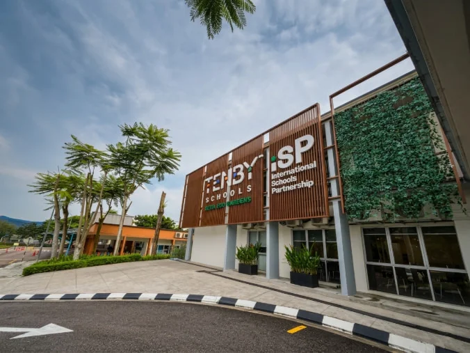 Modern school building with Tenby Schools and ISP logos, surrounded by greenery and a clear sky, viewed from the street.