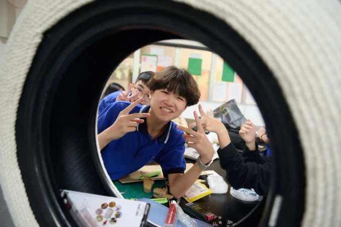 Student in blue shirt smiles and poses with peace signs, framed by a tire, in a classroom setting with colorful posters.