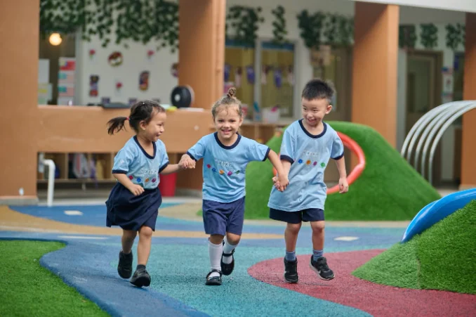 Three children in blue uniforms joyfully run hand in hand on a colorful playground, surrounded by greenery and play structures.