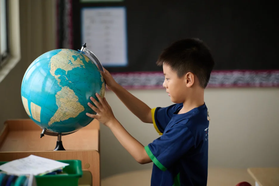 A young boy in a classroom examines a globe, focusing intently on the continents.