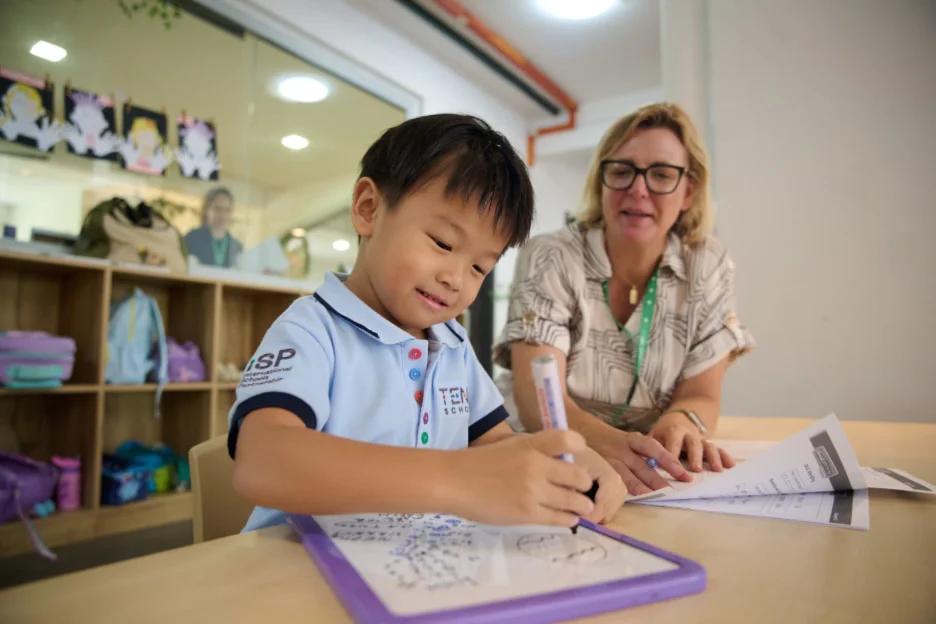 A young boy writes on a whiteboard with a teacher's guidance in a classroom, surrounded by colorful backpacks and shelves.