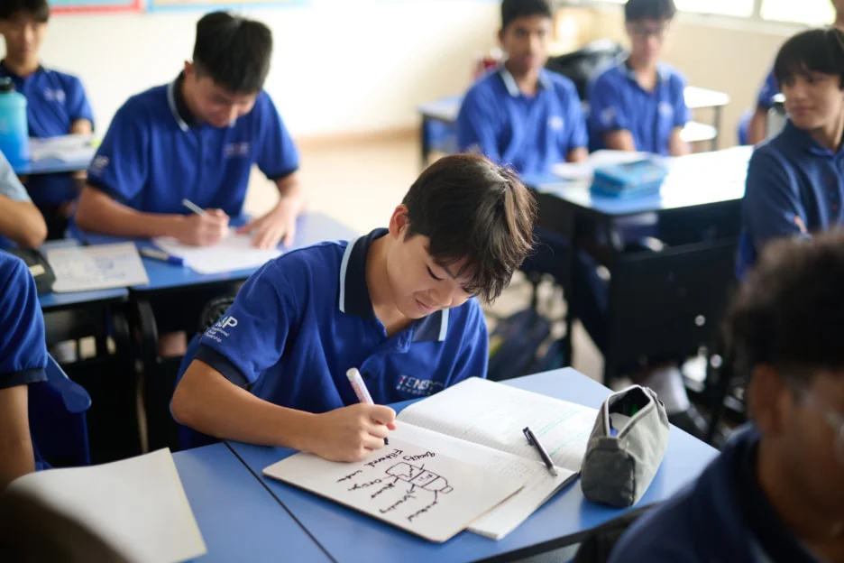 Students in blue uniforms focus on writing in a classroom, with one boy drawing in a notebook at a blue desk.