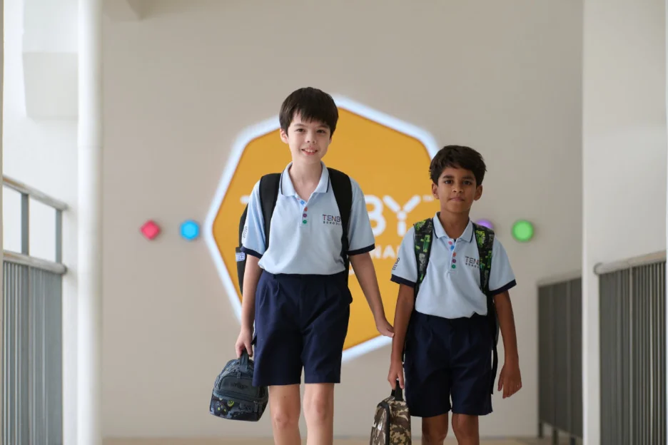Two boys in school uniforms walk down a hallway, smiling and carrying bags, with a colorful sign in the background.