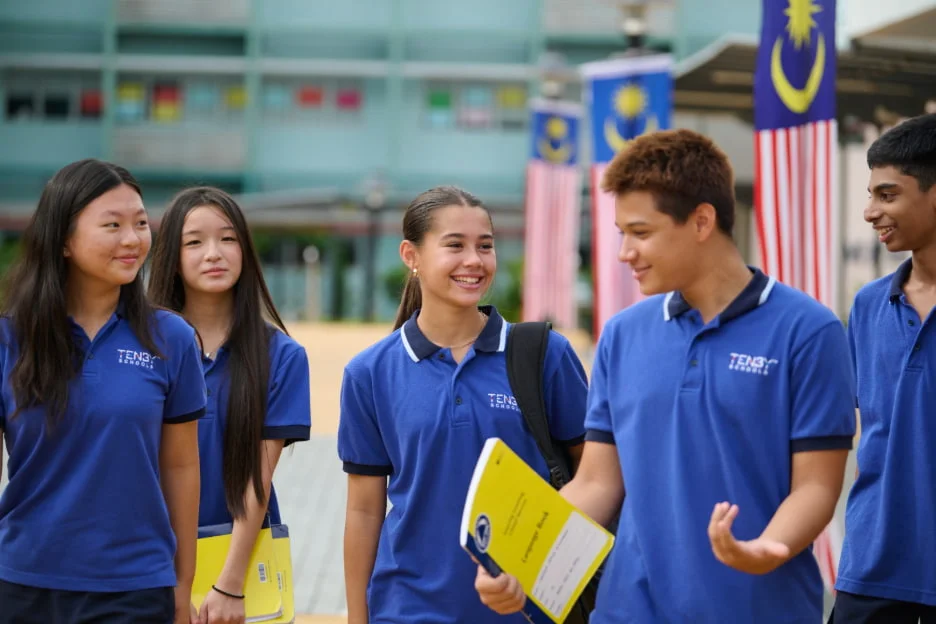 Students in blue uniforms walk and chat, holding notebooks. Malaysian flags hang in the background, suggesting a school setting.