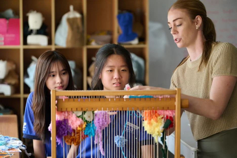 Teacher instructs two students on weaving at a loom, surrounded by colorful yarn in a classroom setting.