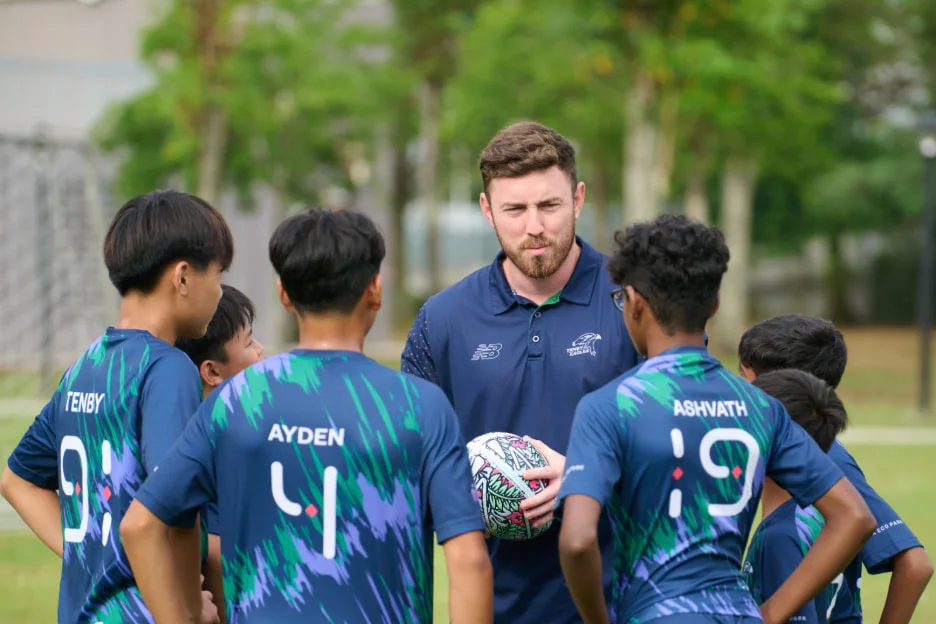 A coach holds a ball, guiding young soccer players in blue jerseys named Ayden, Ashvath, and Tenby during a practice session.